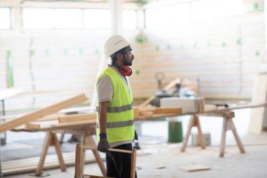 Beyaz miğferi ve güvenlik yeleği olan işçi ahşap bir ev inşa ediyor.