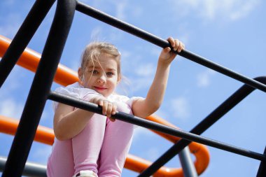 Gülümseyen kız mavi gökyüzünün altında çocuk parkında parmaklıklara tırmanıyor.