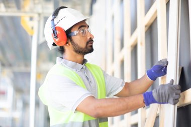 Beyaz miğferi ve güvenlik yeleği olan işçi ahşap bir ev inşa ediyor.