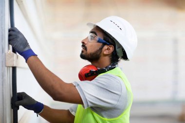 Beyaz miğferi ve güvenlik yeleği olan işçi ahşap bir ev inşa ediyor.