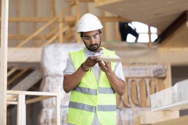 Beyaz miğferi ve güvenlik yeleği olan işçi ahşap bir ev inşa ediyor.