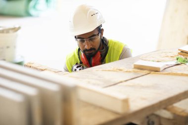 Beyaz miğferi ve güvenlik yeleği olan işçi ahşap bir ev inşa ediyor.
