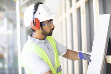 Beyaz miğferi ve güvenlik yeleği olan işçi ahşap bir ev inşa ediyor.
