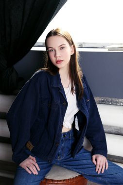 Penceresi ve siyah arka planı olan genç bir kadın model oturuyor.