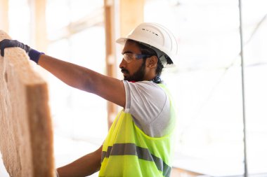Beyaz miğferi ve güvenlik yeleği olan işçi ahşap bir ev inşa ediyor.
