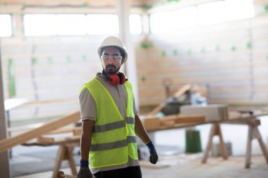 Beyaz miğferi ve güvenlik yeleği olan işçi ahşap bir ev inşa ediyor.