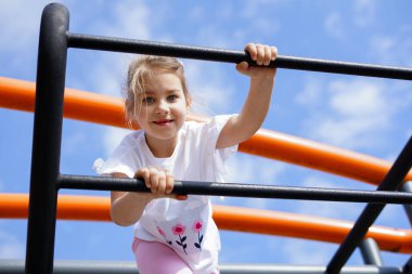 Gülümseyen kız mavi gökyüzünün altında çocuk parkında parmaklıklara tırmanıyor.