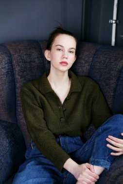 Kot pantolon ve süveter giyen genç bir kadın model koltukta oturuyor.