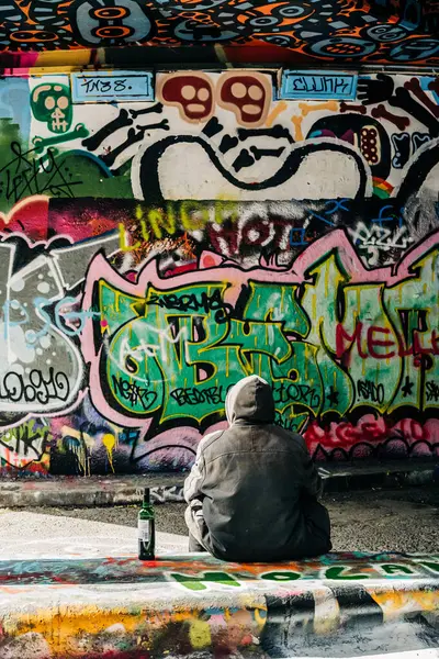 Kapüşonlu adam duvarın yanında elinde şişeyle tek başına oturuyor.