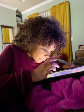 Çocuk gece yatakta yatarken dikkatlice tablet kullanıyor.