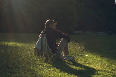 Güneşli bir parkta sırt çantasıyla çimenlerde oturan bir adam.