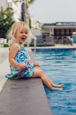 Sarışın kız havuza oturur, neşeyle ayaklarını suya daldırır.