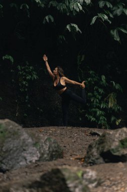 Yoga yapan kadın, kayaların ve yağmur ormanlarının yakınında poz veriyor.