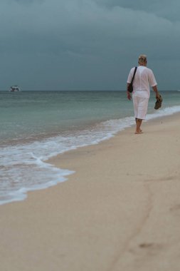 Tropikal plajda sandalet tutan bir adam yalınayak yürüyor.