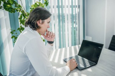 Ofiste dizüstü bilgisayarda çalışan adam, düşünceli bir poz. Modern iç mimari.