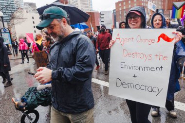 Gülümseyen bir kadın protesto yürüyüşünde Trump karşıtı bir tabela tutuyor.