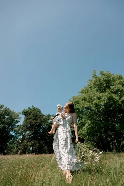 Çıplak ayaklı anne çayırda bebeği ve kır çiçeklerini taşıyor.