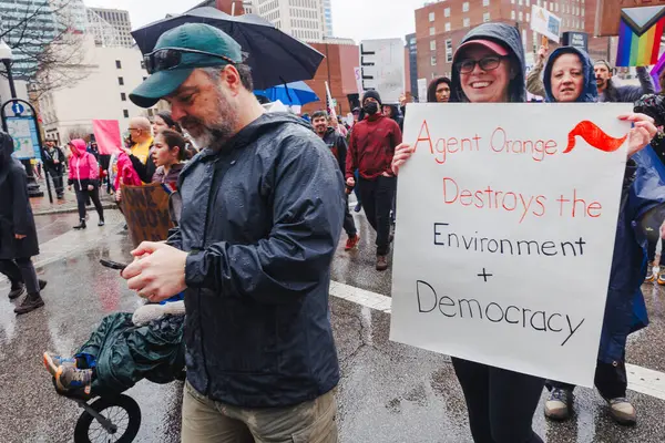 Gülümseyen bir kadın protesto yürüyüşünde Trump karşıtı bir tabela tutuyor.