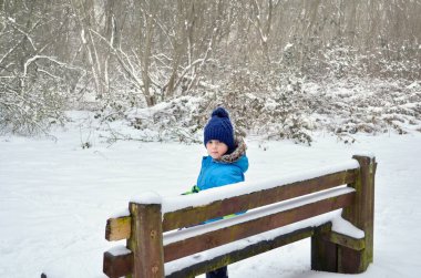 Küçük çocuk bankta oturuyor ve kar parkına bakıyor..