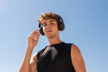 Kulaklıklı genç bir sporcu mavi gökyüzü arkaplanından kameraya bakıyor. 