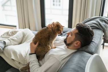 Yan gören adam evdeki kanepede uzanırken kediyi göğsünden okşuyor.