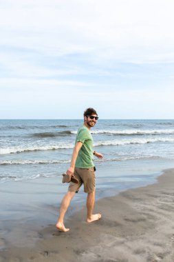 Sahilde yürüyen bir adam kameraya gülümsüyor.