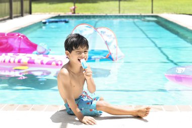 Tişörtsüz çocuk yaz güneşinde havuz kenarında otururken buzlu dondurma yiyor.