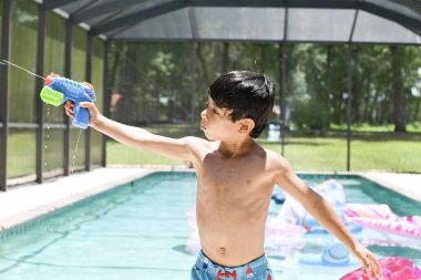 Tişörtsüz çocuk arka bahçede oynarken su tabancasını doğrultuyor..