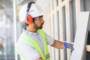 Miğferli ve ceketli işçi ahşap bir salonda çalışıyor.