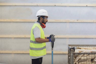 Miğferli ve ceketli işçi ahşap bir salonda çalışıyor.