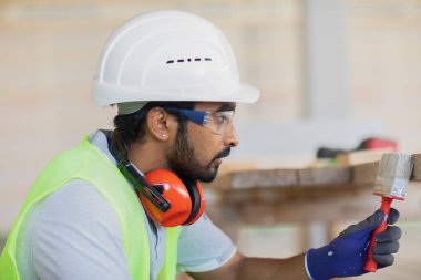 Kask ve güvenlik ceketli işçi ahşap salonda çalışıyor.