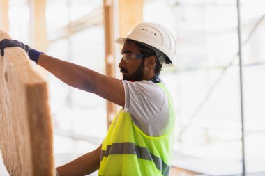 Miğferli ve ceketli işçi ahşap bir salonda çalışıyor.