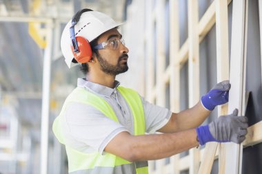 Miğferli ve ceketli işçi ahşap bir salonda çalışıyor.