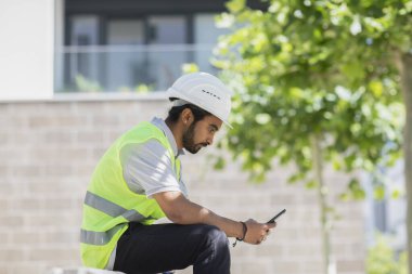 Kasklı ve güvenlik önlüklü işçi dışarıdaki tableti kontrol ediyor.