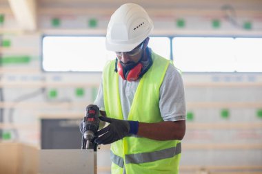 Kask ve güvenlik ceketli işçi ahşap salonda çalışıyor.