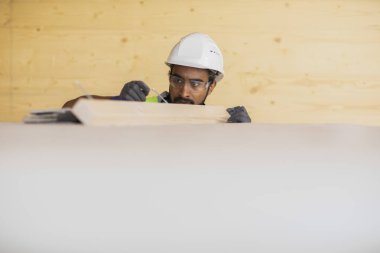 Miğferli ve ceketli işçi ahşap bir salonda çalışıyor.