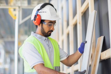 Miğferli ve ceketli işçi ahşap bir salonda çalışıyor.