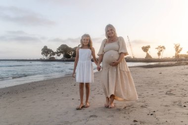 Anne ve kızı gün batımında kumsalda el ele tutuşuyorlar.