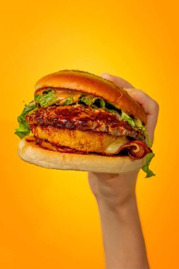 Turuncu bir arka planda parlak sulu bir burger tutuyor.