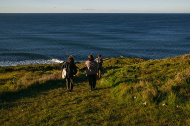 Tourists walking down green hill towards ocean on sunny day