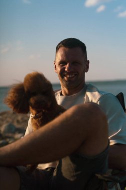 Adam gün batımında köpekle otururken gülümsüyor.