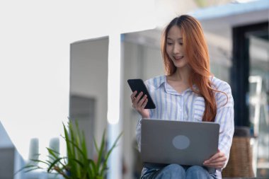 Kadın bir sandalyede oturuyor ve dizüstü bilgisayar ve cep telefonu kullanıyor.