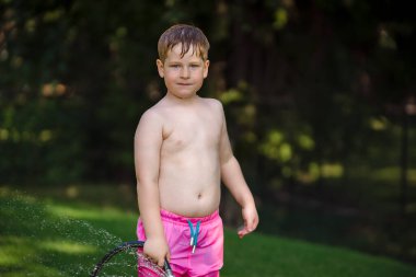 Arka bahçede su hortumuyla oynayan gömleksiz çocuk.