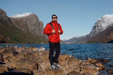 Norveç 'teki fiyortta taşların üzerinde tek başına duran kırmızı ceketli sakallı adam.