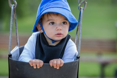 Mavi şapkalı çocuk salıncakta oturuyor, yan tarafa bakıyor.