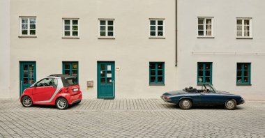 Beyaz bir binanın önüne park edilmiş kırmızı ve mavi üstü açık arabalar.