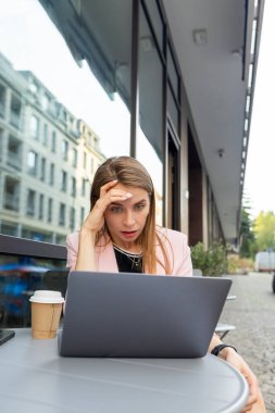 Şaşırmış bir kadın kafenin terasında dizüstü bilgisayara bakıyor.