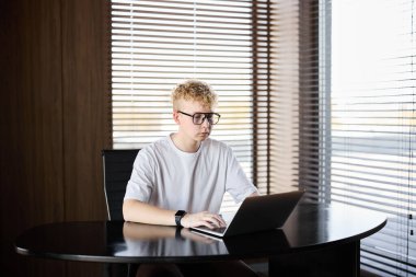 Genç adam aydınlık odadaki modern bir masada dizüstü bilgisayarla çalışıyor.