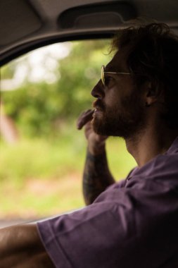 Sakallı, güneş gözlüklü adam, düşünceli bir şekilde arabanın camından dışarı bakıyor.