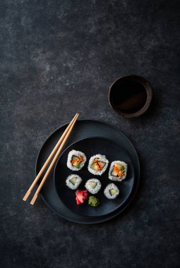 Overhead view plate of sushi rolls and chopsticks on black background.
